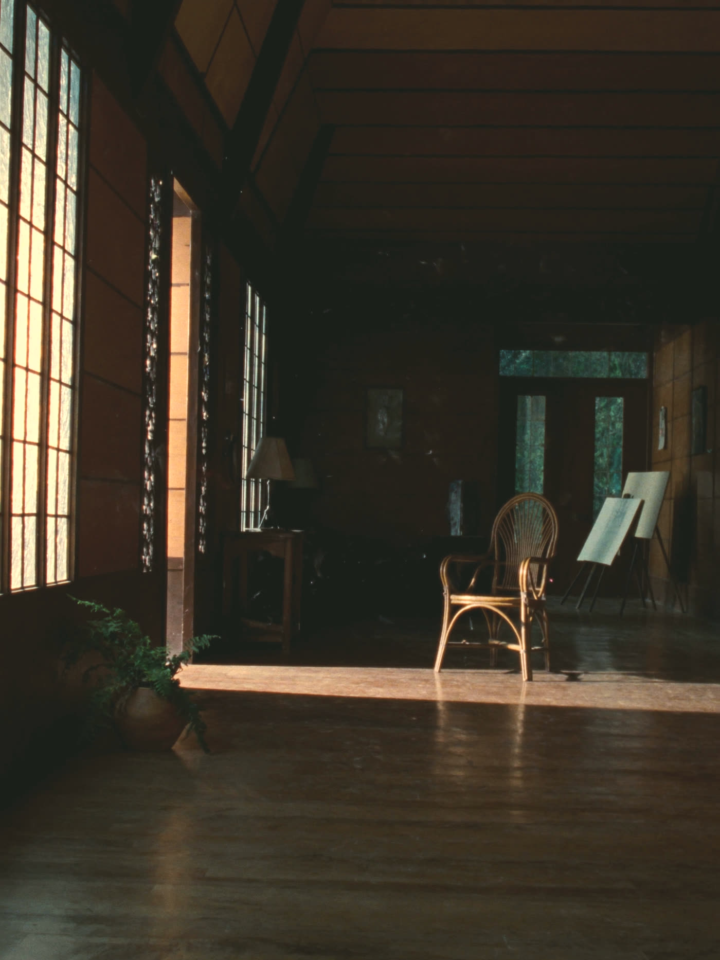 A Filipino home at first light: morning sun falling through capiz shell windows onto a wooden floor, an empty rattan chair at the edge of the room.